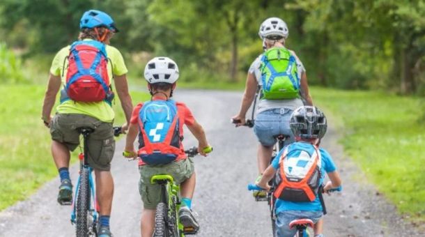 Four people (two adults and two children) ride bicycles on a rural path, wearing helmets and colorful backpacks.