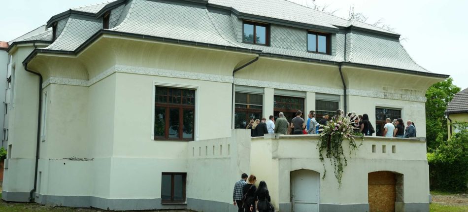 Group of people gathered on a balcony in front of a cream-colored house with a gray tiled roof and decorative molding.