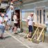 Group of volunteers painting the exterior wall of a building on a sunny day; one person on a ladder applying paint while others work nearby.