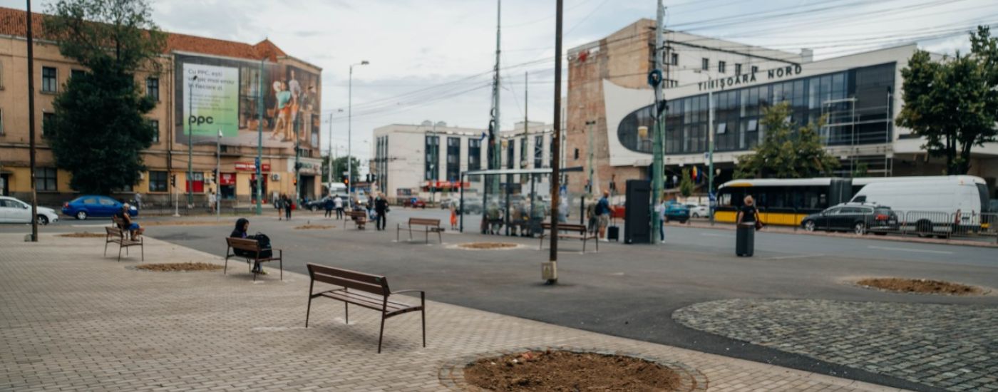 Public square with wooden benches, a dirt circular planting bed, and a street with buses and pedestrians.
