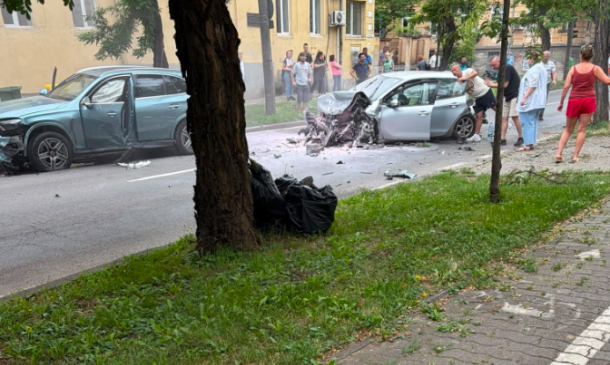 Two cars damaged in a street collision with debris on the road and a crowd of bystanders watching nearby on a residential street.