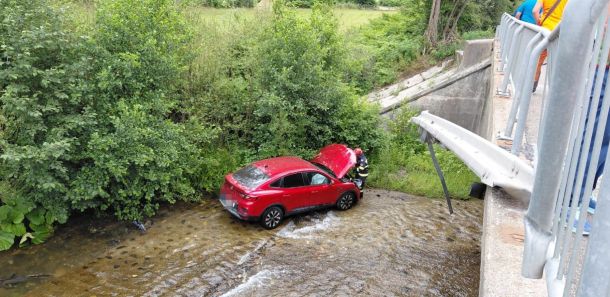 Red SUV sits in a shallow stream beneath a bridge as a rescuer attends to the driver, with onlookers leaning over the railing nearby.
