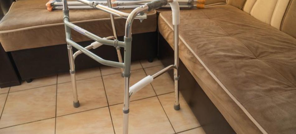 Folding aluminum walker with rubber tips set up in a tiled living room beside a beige sectional sofa.
