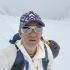 Man in a white jacket takes a selfie in a snowy, mountainous landscape wearing a purple-green cap and white sunglasses with a backpack behind him.