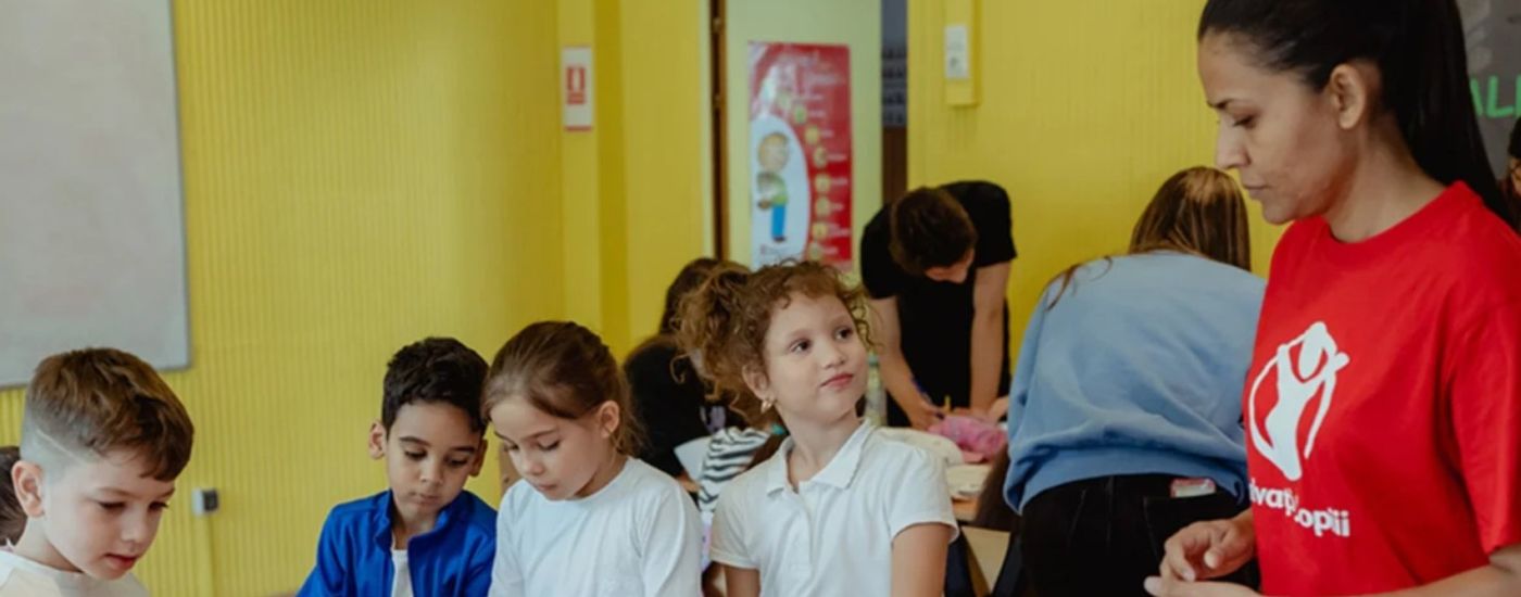 Children participate in a summer camp activity in a yellow classroom; a staff member in a red shirt supervises.