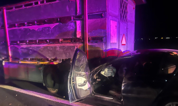 Nighttime crash scene: a black car has collided with the side of a large trailer truck, the driver's door is open and interior is visible with purple emergency lights.