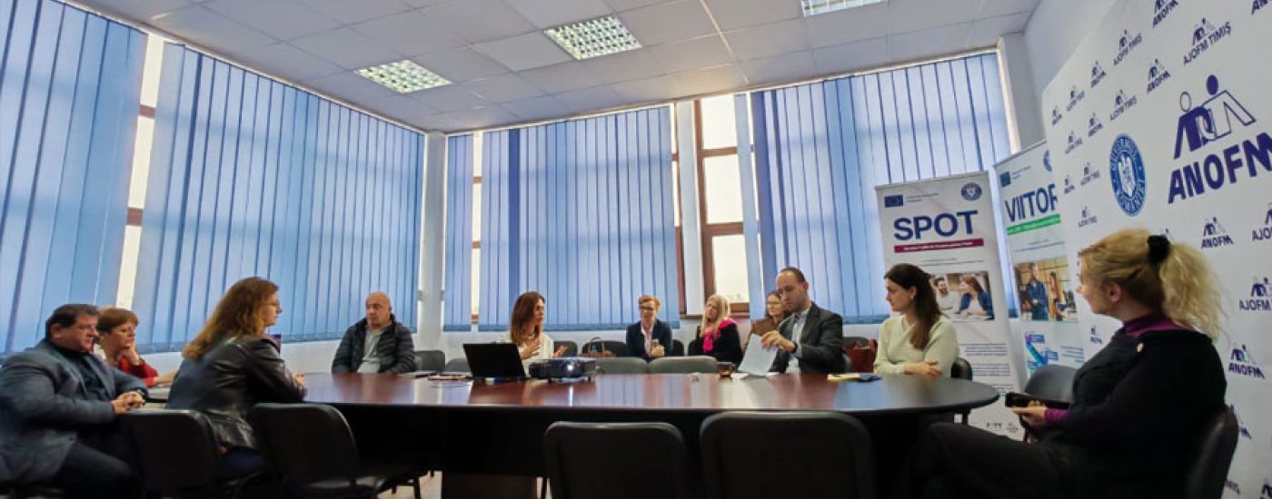 People sit around a large conference table in a meeting room with blue blinds and banners along the wall