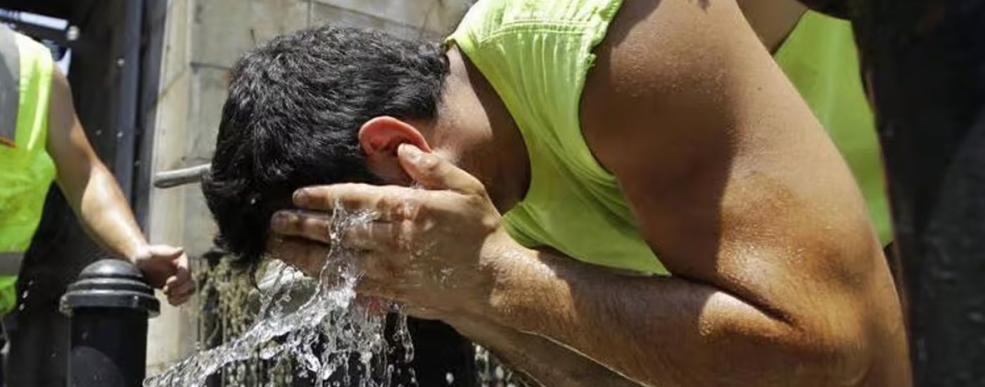 Man in a neon sleeveless shirt splashes water on his head from his hands at an outdoor water fountain, with a coworker in a safety vest nearby.