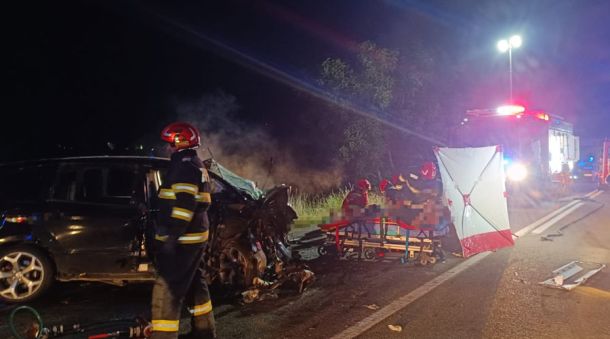 Night-time car crash scene with firefighters and a damaged black sedan on a highway, stretcher and medical crew ready for transport, emergency lights visible in the background
