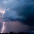 Lightning bolt streaks down through dark storm clouds over a flat horizon at dusk.
