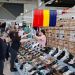 Shoppers browse a busy shoe stall inside a store; rows of sandals and sneakers line the displays with stacked shoeboxes in the background.