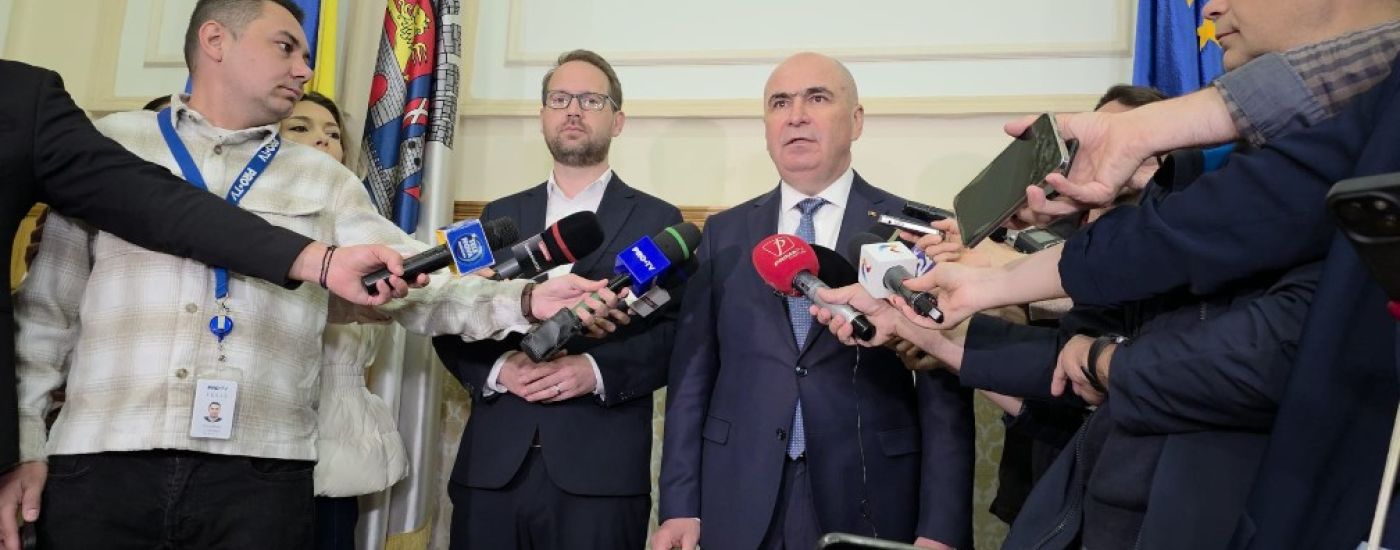 Bald man in a suit addressing reporters with microphones at a press conference, journalists surrounding him.