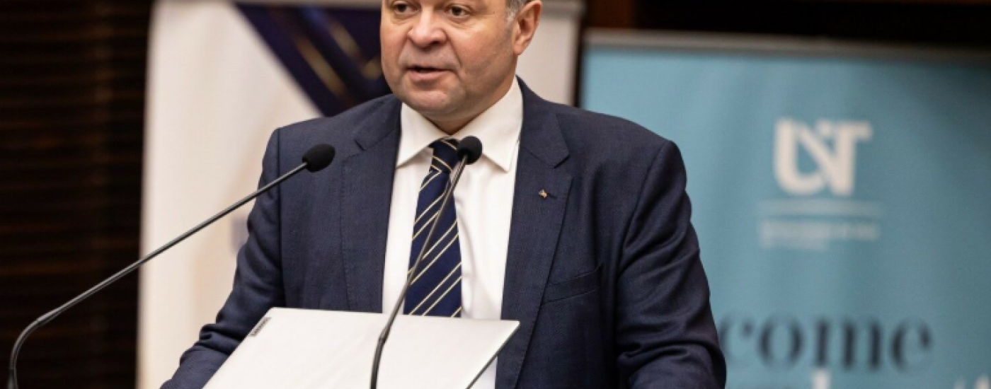 A man in a navy suit and striped tie speaks at a clear podium with two microphones, papers in front, in a formal event setting with banners in the background.