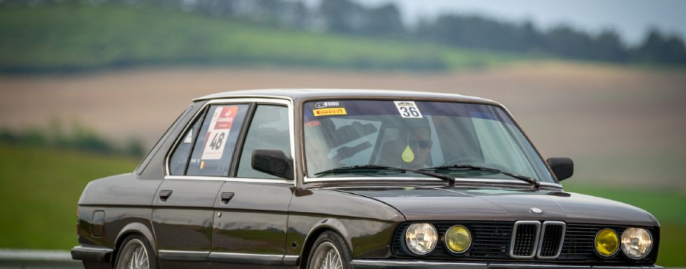 Brown 1980s BMW race car with numbered stickers on a track, front three-quarter view.