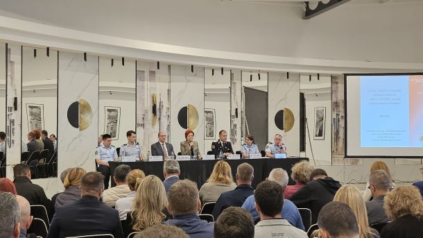 Panel of speakers seated at a long table on a stage, with nameplates and microphones, addressing a seated audience in a conference room.