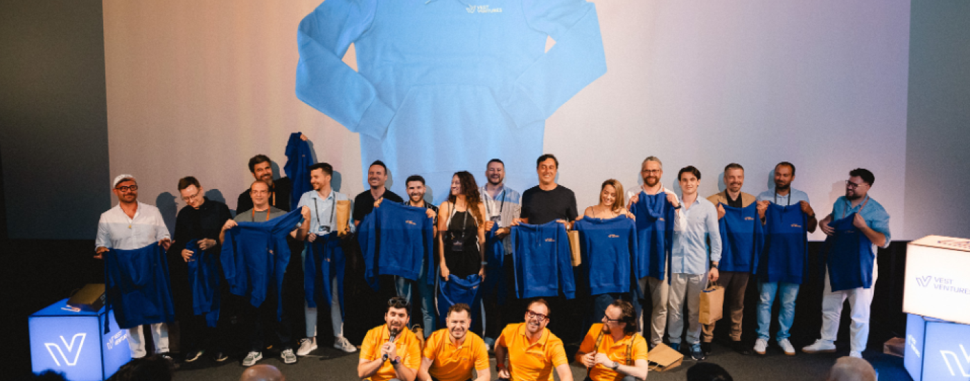 Group of people on a stage posing with blue jackets, in front of a large projected image of a blue hoodie on the screen behind them, an event photo showing staff and speakers holding up jackets.