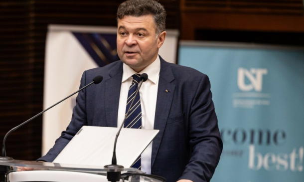 A man in a navy suit and striped tie speaks at a clear podium with two microphones, papers in front, in a formal event setting with banners in the background.