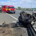 Emergency responders and a red ambulance on a highway beside a severely damaged overturned car and debris on the road flank a crash scene.