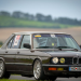 Brown 1980s BMW race car with numbered stickers on a track, front three-quarter view.