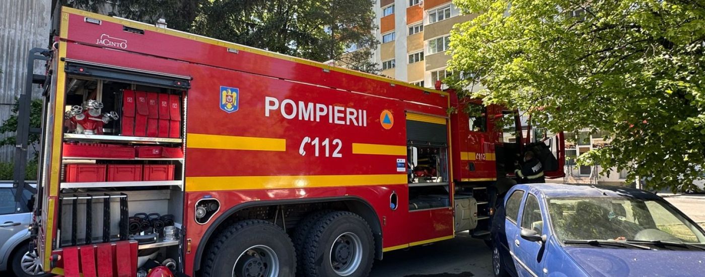 Red fire truck with yellow stripes labeled POMPIERII 112 parked on a city street beside a blue car with a residential building in the background and trees nearby.