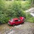 Red SUV sits in a shallow stream beneath a bridge as a rescuer attends to the driver, with onlookers leaning over the railing nearby.