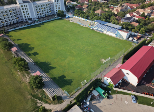 Aerial view of a green rectangular soccer field with goal nets, surrounded by bleachers and buildings. Nearby houses line the hillside beyond the stadium.