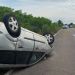 White SUV flipped onto its roof on a rural two-lane road; emergency responders and traffic cones visible in the distance.