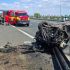 Emergency responders and a red ambulance on a highway beside a severely damaged overturned car and debris on the road flank a crash scene.