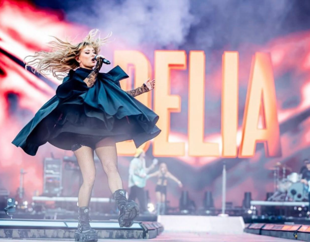 Female singer mid-air on a stage performing with a microphone, wearing a dark blue dress/coat, with large orange letters in the background.