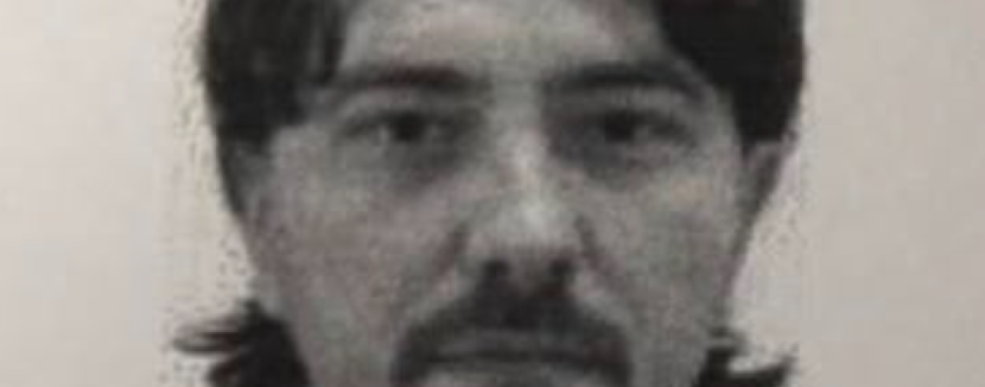 Black-and-white headshot of a man with medium-length dark hair and a goatee, wearing a dark collared shirt, looking at the camera.