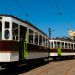 Vintage tram cars parked on cobblestone tracks in a rail yard under a bright blue sky, beside a museum depot with green doors.