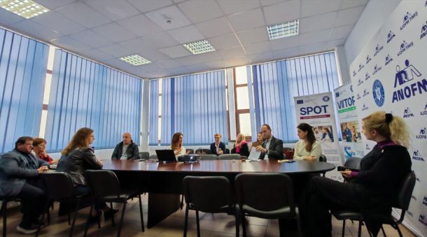 People sit around a large conference table in a meeting room with blue blinds and banners along the wall