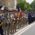 Line of soldiers in uniform standing at attention during a parade, one holding a blue-yellow-red flag.