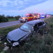 Gray sedan crashed off a rural road into tall weeds, front end damaged, with emergency vehicles and responders arriving in the background at dusk.