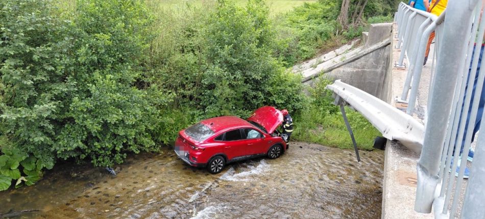 Red SUV sits in a shallow stream beneath a bridge as a rescuer attends to the driver, with onlookers leaning over the railing nearby.