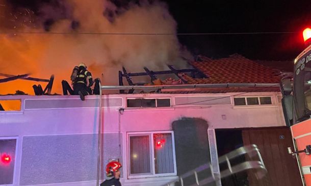 Firefighters on a burning building roof at night as flames and thick smoke rise, with a fire engine in view at the right. (Informative)