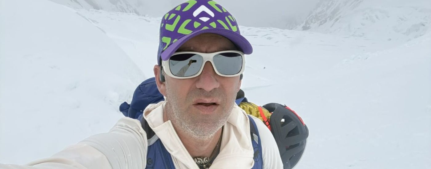 Man in a white jacket takes a selfie in a snowy, mountainous landscape wearing a purple-green cap and white sunglasses with a backpack behind him.