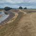 Dirt road along a sloped, rocky riverbank with calm water to the left and distant trees under a cloudy sky.