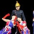 Actors in Union Jack costumes perform on stage, two women posing dramatically while a man in a firefighter helmet stands behind them against a black background