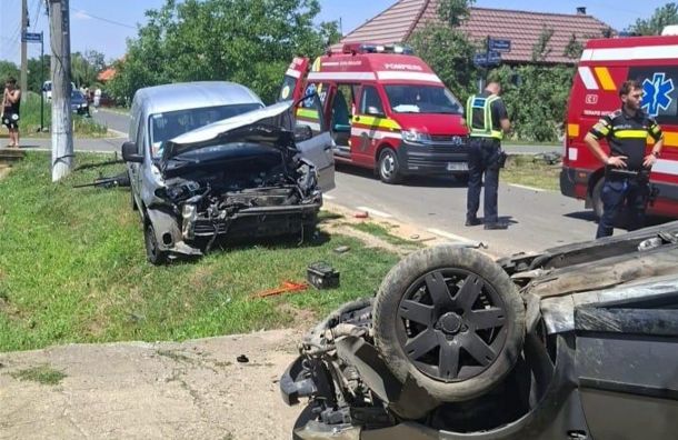 Two damaged vehicles after a crash: a silver van with severe front-end damage on the grass and a flipped car on the road, with paramedics and ambulances nearby on a sunny day.