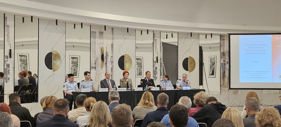 Panel of speakers seated at a long table on a stage, with nameplates and microphones, addressing a seated audience in a conference room.
