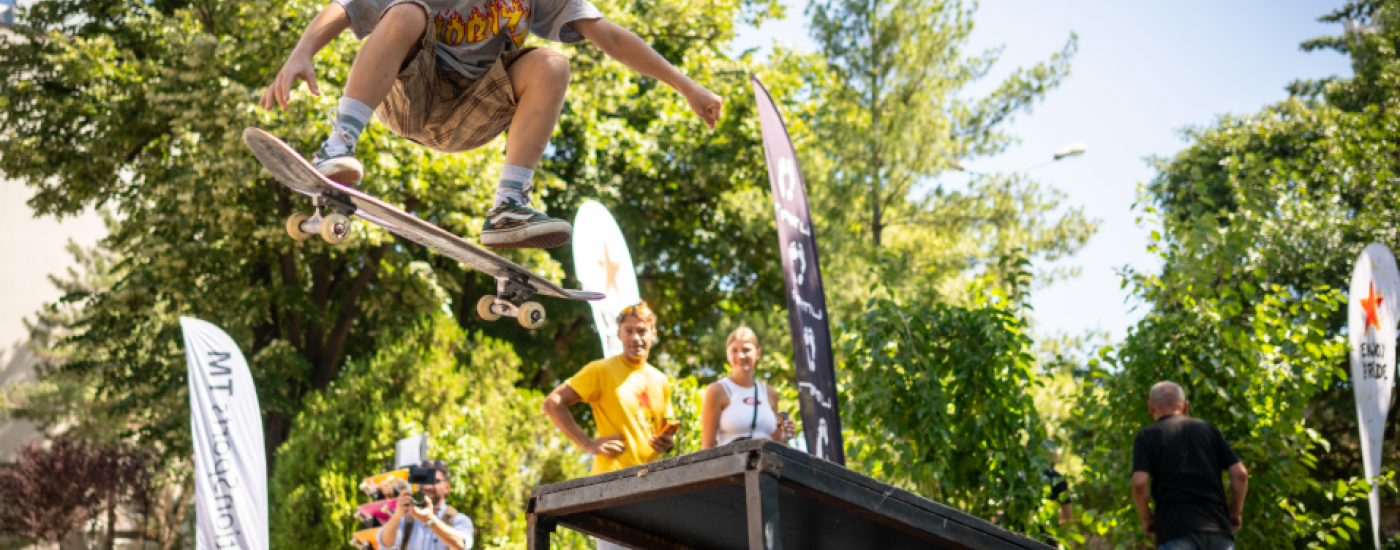 Skater launches off a ramp, mid-air, performing a kickflip while others watch in a sunny park.