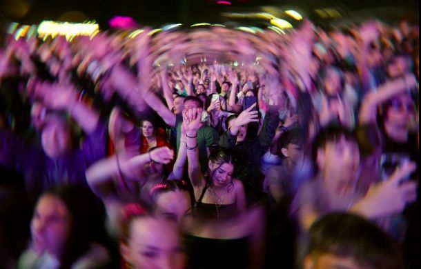 Large crowd at a concert with hands raised and phones recording, colorful stage lights glow in the background.