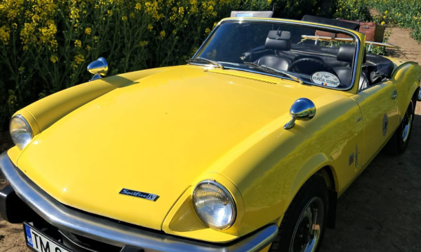 Yellow vintage convertible (Triumph Spitfire) parked on a rural road beside a canola field in bloom on a sunny day.