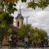 Group of cyclists riding along a tree-lined street with a church steeple visible in the background.