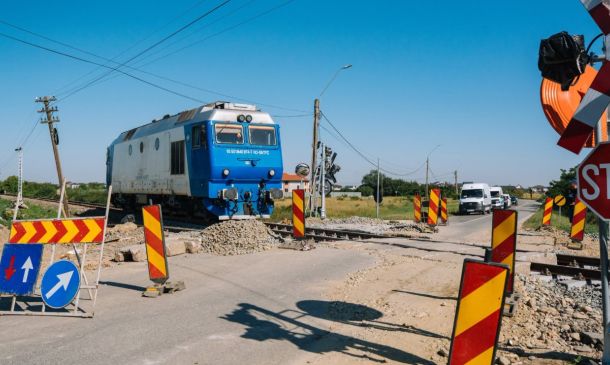 Blue locomotive at a rural railroad crossing under construction, with orange-yellow striped barriers and warning signs nearby.