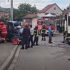 Firefighters and responders work at a car crash on a street with a tow truck and debris nearby, a fire truck in the background.