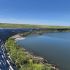 Curved wall of stacked concrete bags along a calm water body under a clear blue sky and grassy embankment beyond.