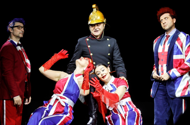 Actors in Union Jack costumes perform on stage, two women posing dramatically while a man in a firefighter helmet stands behind them against a black background