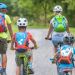 Four people (two adults and two children) ride bicycles on a rural path, wearing helmets and colorful backpacks.